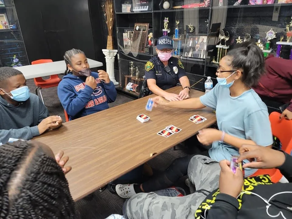students are sitting at the table with a volunteer and a police officer playing uno cards.